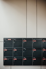A set of sixteen black mailboxes arranged in a four by four grid is mounted on a light-colored wall. Each mailbox has a label with red accents that indicate unit numbers. The overall setting is minimal and organized.