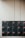 A set of sixteen black mailboxes arranged in a four by four grid is mounted on a light-colored wall. Each mailbox has a label with red accents that indicate unit numbers. The overall setting is minimal and organized.