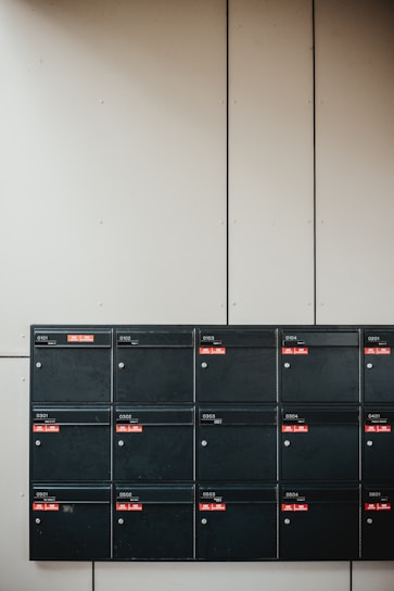 A set of sixteen black mailboxes arranged in a four by four grid is mounted on a light-colored wall. Each mailbox has a label with red accents that indicate unit numbers. The overall setting is minimal and organized.