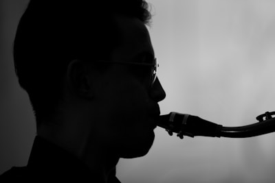A silhouette of a person playing a saxophone. The image captures the side profile, highlighting the curve of the instrument and the outline of the individual's face and hair. The background is a soft gradient, adding a calm atmosphere to the composition.