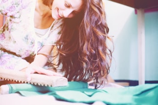 A woman with long, flowing brown hair smiles joyfully while engaging in a hands-on activity using a wooden tool on a flat surface. She is wearing a floral-patterned outfit and the setting has soft, pastel colors.