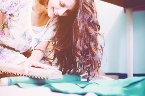 A woman with long, flowing brown hair smiles joyfully while engaging in a hands-on activity using a wooden tool on a flat surface. She is wearing a floral-patterned outfit and the setting has soft, pastel colors.