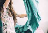 A person with long hair is holding and adjusting a large piece of teal fabric, possibly a bedsheet. They are wearing a floral patterned garment, and the setting suggests a domestic environment.