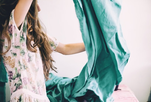 A person with long hair is holding and adjusting a large piece of teal fabric, possibly a bedsheet. They are wearing a floral patterned garment, and the setting suggests a domestic environment.