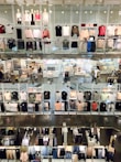 A multi-level clothing store interior featuring numerous shelves and mannequins displaying various garments. Clothes of different colors, including shirts, sweaters, and jackets, are arranged neatly. The store has bright lighting, and there are shoppers visible in some sections.