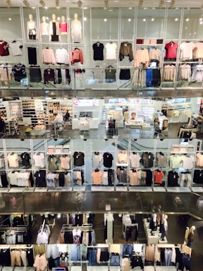 A multi-level clothing store interior featuring numerous shelves and mannequins displaying various garments. Clothes of different colors, including shirts, sweaters, and jackets, are arranged neatly. The store has bright lighting, and there are shoppers visible in some sections.
