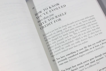 An open book is displayed with a visible page featuring large bold text at the top reading 'HOW TO KNOW YOU'VE EVOLVED more than you GIVE YOURSELF CREDIT FOR.' Below is a paragraph discussing personal growth and self-reflection, encouraging readers to acknowledge their achievements.