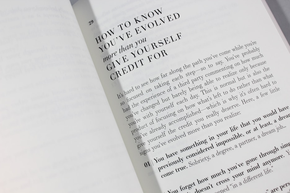 An open book is displayed with a visible page featuring large bold text at the top reading 'HOW TO KNOW YOU'VE EVOLVED more than you GIVE YOURSELF CREDIT FOR.' Below is a paragraph discussing personal growth and self-reflection, encouraging readers to acknowledge their achievements.