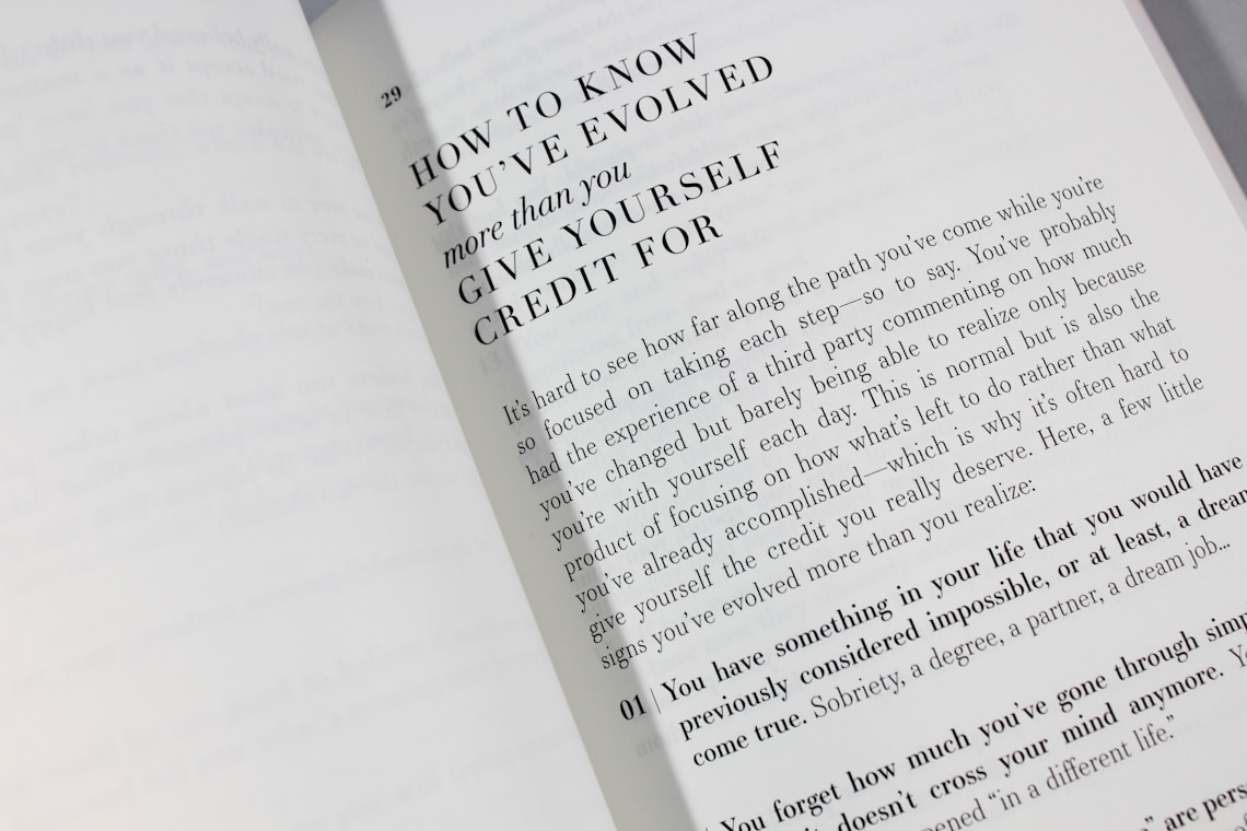 An open book is displayed with a visible page featuring large bold text at the top reading 'HOW TO KNOW YOU'VE EVOLVED more than you GIVE YOURSELF CREDIT FOR.' Below is a paragraph discussing personal growth and self-reflection, encouraging readers to acknowledge their achievements.