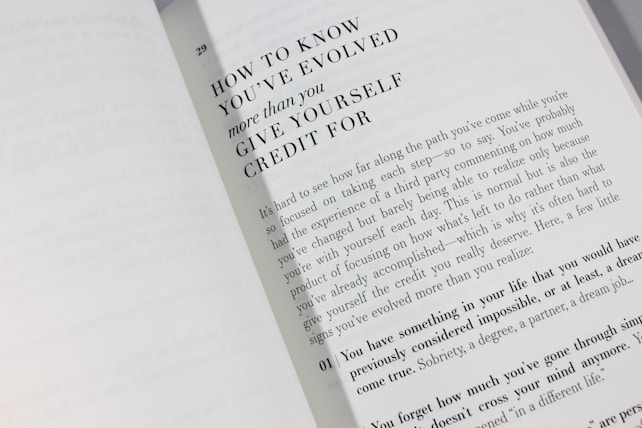 An open book is displayed with a visible page featuring large bold text at the top reading 'HOW TO KNOW YOU'VE EVOLVED more than you GIVE YOURSELF CREDIT FOR.' Below is a paragraph discussing personal growth and self-reflection, encouraging readers to acknowledge their achievements.