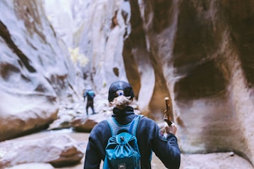 A person with a ponytail, wearing a blue backpack and hat, holds a walking stick while hiking through a narrow canyon. The walls of the canyon are tall and rugged, creating a dramatic natural corridor. Another hiker is visible in the distance, emphasizing the scale of the landscape.