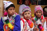 Three boys are wearing traditional, colorful attire with intricate patterns and designs. They don vibrant, multi-colored hats and ponchos with tassels. In the background, there is a woman and some framed artwork. The setting appears to be cultural or festival-driven.