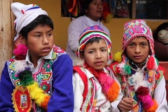 Three boys are wearing traditional, colorful attire with intricate patterns and designs. They don vibrant, multi-colored hats and ponchos with tassels. In the background, there is a woman and some framed artwork. The setting appears to be cultural or festival-driven.
