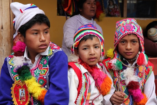 Three boys are wearing traditional, colorful attire with intricate patterns and designs. They don vibrant, multi-colored hats and ponchos with tassels. In the background, there is a woman and some framed artwork. The setting appears to be cultural or festival-driven.