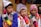 Three boys are wearing traditional, colorful attire with intricate patterns and designs. They don vibrant, multi-colored hats and ponchos with tassels. In the background, there is a woman and some framed artwork. The setting appears to be cultural or festival-driven.