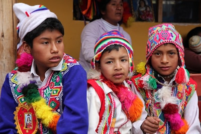 Three boys are wearing traditional, colorful attire with intricate patterns and designs. They don vibrant, multi-colored hats and ponchos with tassels. In the background, there is a woman and some framed artwork. The setting appears to be cultural or festival-driven.