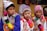 Three boys are wearing traditional, colorful attire with intricate patterns and designs. They don vibrant, multi-colored hats and ponchos with tassels. In the background, there is a woman and some framed artwork. The setting appears to be cultural or festival-driven.