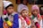Three boys are wearing traditional, colorful attire with intricate patterns and designs. They don vibrant, multi-colored hats and ponchos with tassels. In the background, there is a woman and some framed artwork. The setting appears to be cultural or festival-driven.