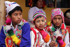 Three boys are wearing traditional, colorful attire with intricate patterns and designs. They don vibrant, multi-colored hats and ponchos with tassels. In the background, there is a woman and some framed artwork. The setting appears to be cultural or festival-driven.