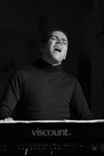 A person appears to be passionately playing a keyboard, possibly a musician engaged in a performance. The expression on their face suggests deep emotion or concentration. The setting is dimly lit, enhancing the focus on the individual's expression.