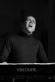 A person appears to be passionately playing a keyboard, possibly a musician engaged in a performance. The expression on their face suggests deep emotion or concentration. The setting is dimly lit, enhancing the focus on the individual's expression.