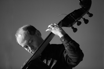 A black and white image of a person playing a double bass, focusing intently on the instrument. The lighting creates strong contrasts, emphasizing the musician's concentration and the texture of the instrument.