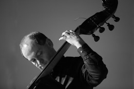 A black and white image of a person playing a double bass, focusing intently on the instrument. The lighting creates strong contrasts, emphasizing the musician's concentration and the texture of the instrument.