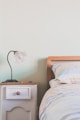 A cozy bedroom interior with a wooden headboard visible on the right side of the image. A bedside table on the left is topped with a decorative lamp, featuring a white, petal-like lampshade. The table, painted in a soft white, has one drawer with a round knob. The bedding on the bed features a striped pattern with muted colors.