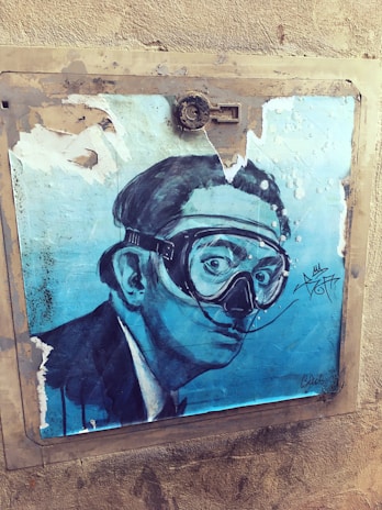 A mural artwork featuring a monochromatic blue portrait of a man wearing a snorkel mask. The background is textured with visible peeling, giving a distressed appearance. The artwork includes stylized signatures and is framed in a dull beige border mounted on a rough, beige wall.