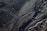 Dark sand with intricate patterns formed by the movement of water, creating a series of wavy lines and textures. Small stones and debris are scattered across the surface, catching the light.