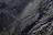 Dark sand with intricate patterns formed by the movement of water, creating a series of wavy lines and textures. Small stones and debris are scattered across the surface, catching the light.