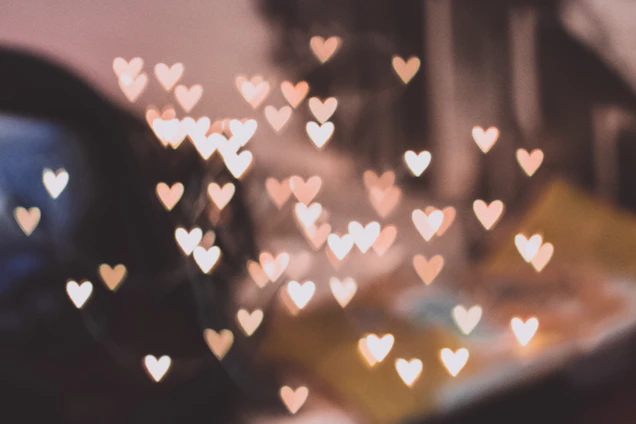 A soft-focus image of glowing rose gold ribbons and floating hearts against a deep burgundy gradient background.