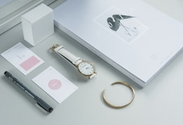 A minimalist arrangement featuring a closed notepad or planner with an elegant cover design, a white strap wristwatch, a golden bracelet, a few minimalistic cards, a black pen, and a white cube object. The color scheme is predominantly light, with a calming and sophisticated aesthetic.