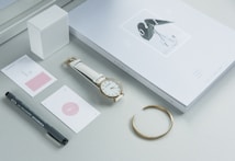A minimalist arrangement featuring a closed notepad or planner with an elegant cover design, a white strap wristwatch, a golden bracelet, a few minimalistic cards, a black pen, and a white cube object. The color scheme is predominantly light, with a calming and sophisticated aesthetic.