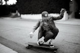 A person is crouching low while riding a skateboard on a quiet street. Wearing a cap, striped shirt, and denim jacket, they exude a casual style. The background includes a sidewalk and some shrubbery, with the scene captured in black and white, giving it a nostalgic feel.