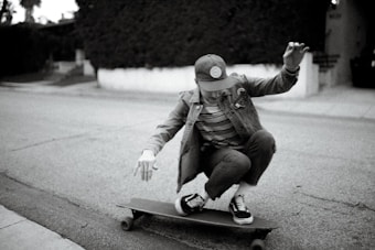 A person is crouching low while riding a skateboard on a quiet street. Wearing a cap, striped shirt, and denim jacket, they exude a casual style. The background includes a sidewalk and some shrubbery, with the scene captured in black and white, giving it a nostalgic feel.