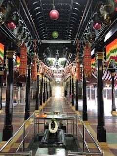 A richly decorated hall with intricately designed multi-colored lanterns hanging from the ceiling along with strings of lights. The hall is lined with polished black pillars featured with gold accents. At the far end of the hall, daylight pours through an open doorway. In the foreground, there is a metallic structure with candles and a wreath.