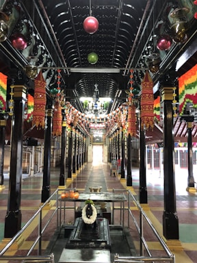 A richly decorated hall with intricately designed multi-colored lanterns hanging from the ceiling along with strings of lights. The hall is lined with polished black pillars featured with gold accents. At the far end of the hall, daylight pours through an open doorway. In the foreground, there is a metallic structure with candles and a wreath.