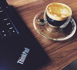 A cup of espresso is placed on a wooden surface beside a laptop with 'ThinkPad' branding. The coffee is in a clear glass cup with a matching saucer, and a small spoon rests beside it.