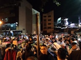 A bustling outdoor night market with numerous people browsing through racks of clothes. Illuminated by artificial lights, the scene is busy and energetic with various signs displaying promotions and prices. The surrounding buildings contribute to the vibrant urban atmosphere.