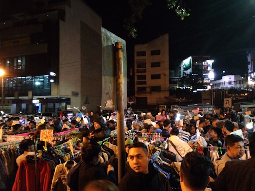 A bustling outdoor night market with numerous people browsing through racks of clothes. Illuminated by artificial lights, the scene is busy and energetic with various signs displaying promotions and prices. The surrounding buildings contribute to the vibrant urban atmosphere.