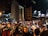 A bustling outdoor night market with numerous people browsing through racks of clothes. Illuminated by artificial lights, the scene is busy and energetic with various signs displaying promotions and prices. The surrounding buildings contribute to the vibrant urban atmosphere.