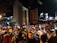 A bustling outdoor night market with numerous people browsing through racks of clothes. Illuminated by artificial lights, the scene is busy and energetic with various signs displaying promotions and prices. The surrounding buildings contribute to the vibrant urban atmosphere.
