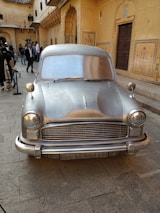 A silver, vintage car is prominently displayed in a courtyard surrounded by ornate, yellow sandstone architecture. Several people are gathered around, some taking photos and others walking by. The building features intricate carvings and archways, characteristic of traditional Indian designs.