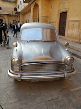 A silver, vintage car is prominently displayed in a courtyard surrounded by ornate, yellow sandstone architecture. Several people are gathered around, some taking photos and others walking by. The building features intricate carvings and archways, characteristic of traditional Indian designs.