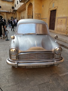 A silver, vintage car is prominently displayed in a courtyard surrounded by ornate, yellow sandstone architecture. Several people are gathered around, some taking photos and others walking by. The building features intricate carvings and archways, characteristic of traditional Indian designs.
