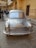 A silver, vintage car is prominently displayed in a courtyard surrounded by ornate, yellow sandstone architecture. Several people are gathered around, some taking photos and others walking by. The building features intricate carvings and archways, characteristic of traditional Indian designs.
