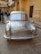 A silver, vintage car is prominently displayed in a courtyard surrounded by ornate, yellow sandstone architecture. Several people are gathered around, some taking photos and others walking by. The building features intricate carvings and archways, characteristic of traditional Indian designs.