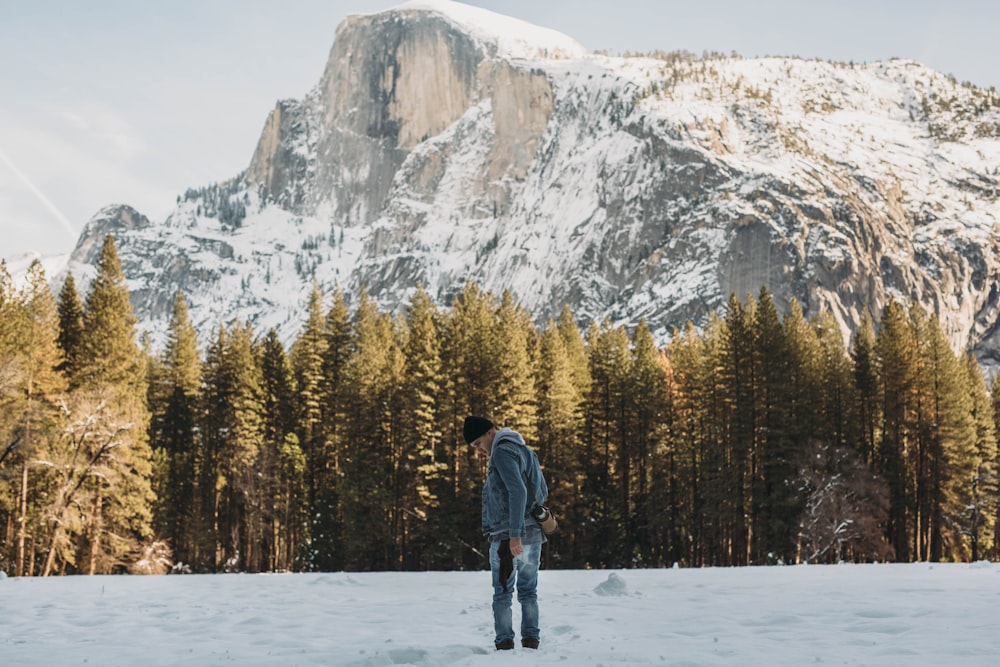 man standing on snow field near trees and mountain during day