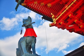 A statue of a fox with a red bib around its neck holds a key in its mouth. The fox is positioned under a structure with intricate wooden beams painted bright red. The sky is a vibrant blue with scattered white clouds.